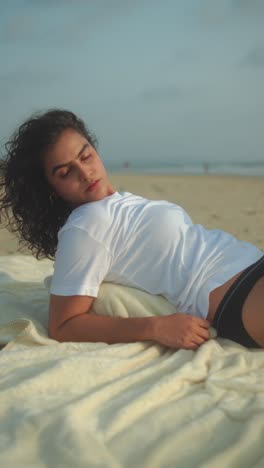 indian woman in white t-shirt and bikini bottoms relaxes on light blanket along sandy beach