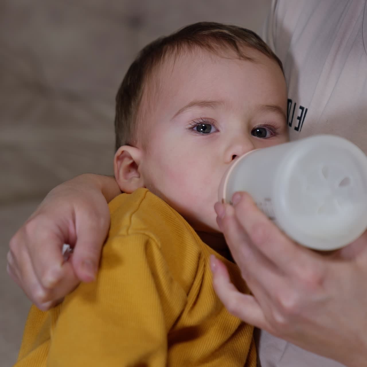 Baby boy lying in mommy hands is fed from a bottle. Kid looks straight ahead while eating. Top view close up