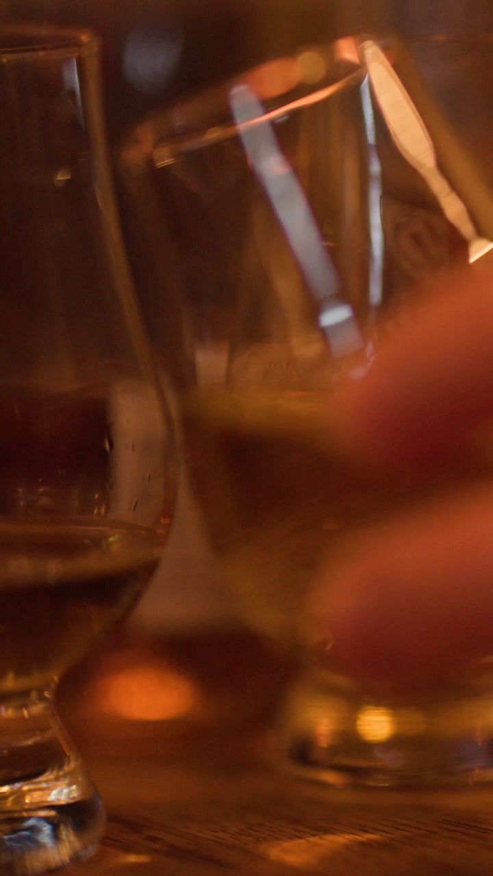 Una mano si protende verso una fila di bicchieri da whisky sotto una calda e suggestiva illuminazione da pub.