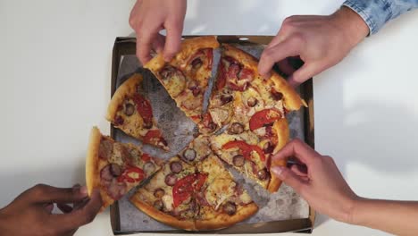 Hands-taking-pizza-cuts-from-paper-box-on-table.-Top-view.-Unrecognizable-people-simultaneously-spreading-arms-to-get-slices-of