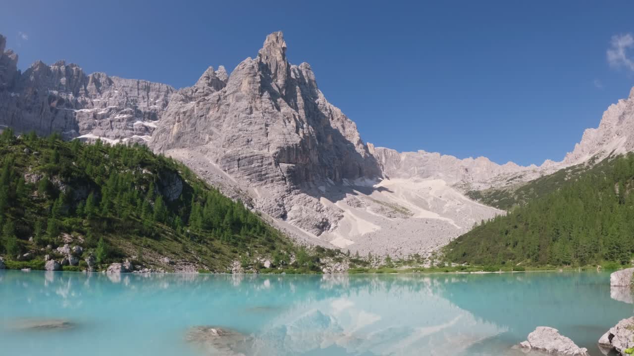 A turquoise lake reflects the towering rocky peaks and green pine-covered slopes surrounding it. The clear blue sky and untouched natural setting create a tranquil and majestic mountain landscape.