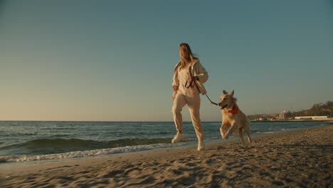 Trotando-Y-Jugando-Con-Sus-Mascotas,-Una-Chica-Rubia-En-La-Playa-Corre-Con-Su-Gran-Perro-Gris-Por-La-Mañana