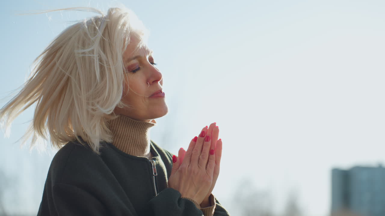 Smiling mature woman with short blonde hair enjoying sunlight outdoors, eyes closed, wearing green jacket and beige turtleneck, red nails clasped together, expressing peace, gratitude in natural setting