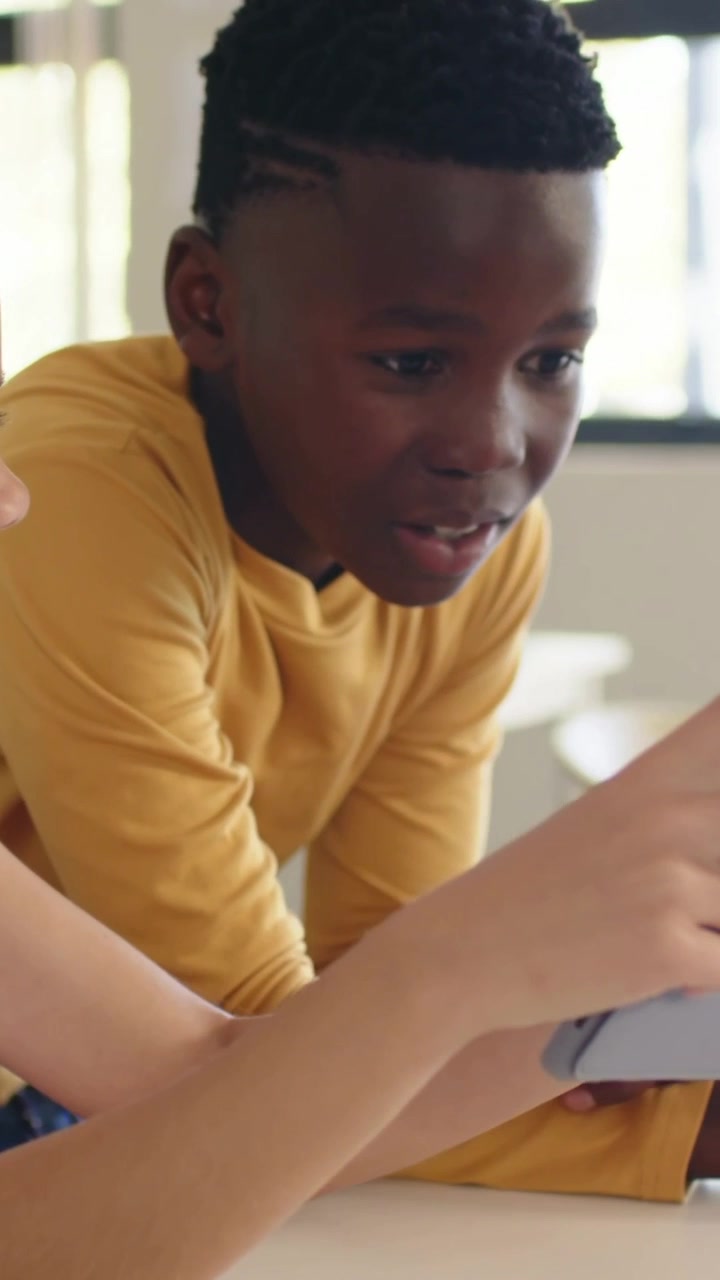 Vidéo verticale : Un enfant lève une tablette grise, un garçon en jaune penché à table est encouragé, partageant du contenu.