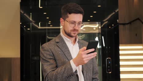 hombre de negocios serio sosteniendo un teléfono inteligente escribiendo un mensaje de pie cerca de un ascensor moderno. gerente inteligente ceo usando aplicaciones móviles de teléfonos celulares. soluciones de aplicaciones de tecnología digital para el desarrollo corporativo de negocios