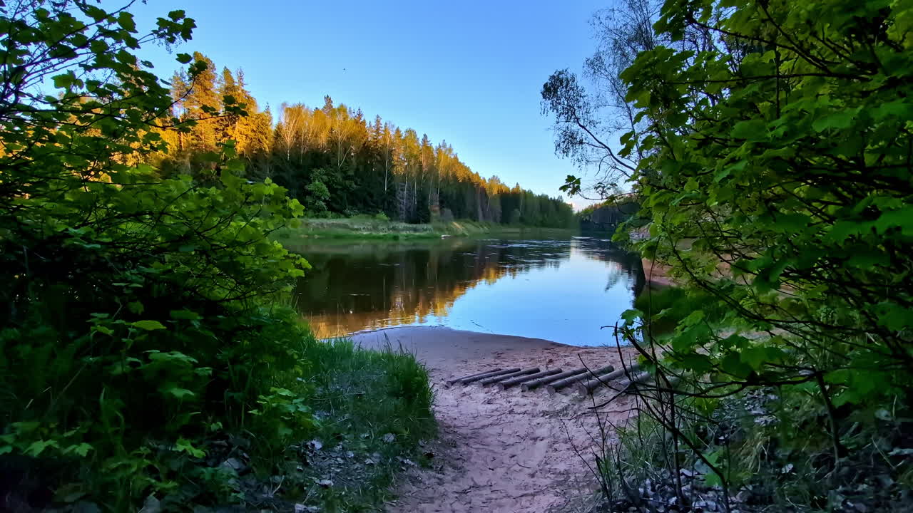 Margem de rio florestal arenosa com árvores douradas refletindo em água calma sob a luz do entardecer.