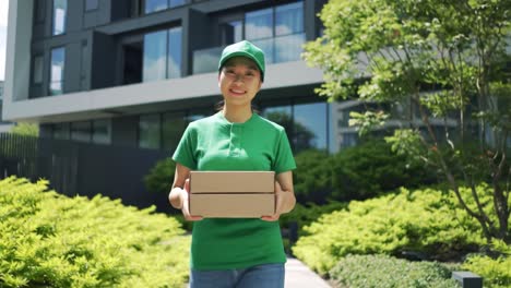 A cheerful delivery person wearing a green shirt and cap carries two boxes towards a contemporary apartment. The sunny day enhances a welcoming atmosphere in the lush surroundings.