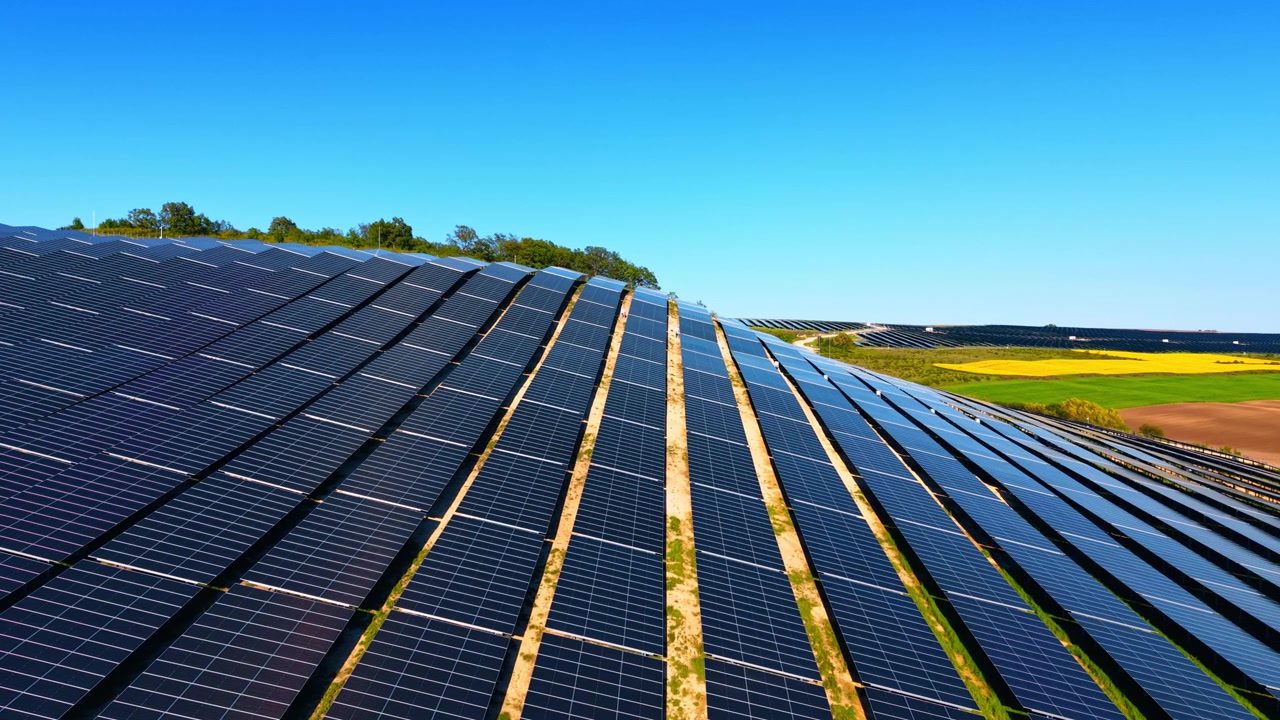 File di pannelli solari su una collina con terreni agricoli rurali. Prospettiva diagonale di schiere fotovoltaiche, sentieri di terra asciutta e campi verdi sotto un cielo limpido.
