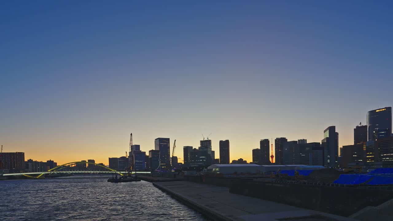 東京のウォーターフロントのスカイラインが、暖色系のオレンジから青への夕暮れの空にシルエットで浮かび上がる。緑のアーチ橋、穏やかな川に浮かぶはしけ、建設クレーン、赤い塔が見える静止広角ショット
