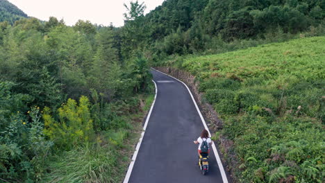 rising drone shot of woman on scooter driving on empty road in middle of bamboo forest in china