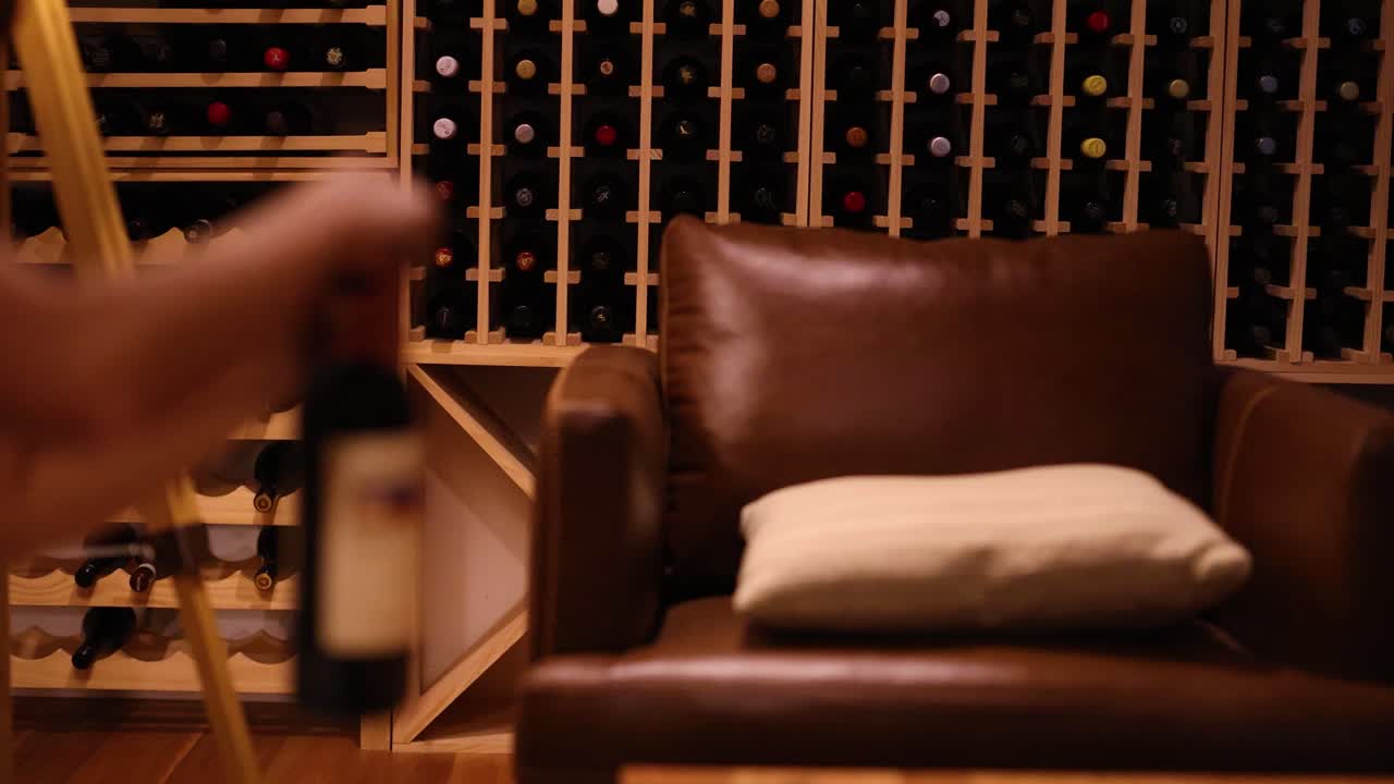 A hand holds a wine bottle in a cozy cellar with a leather chair and wooden racks, under warm lighting