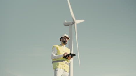 a medium shot of a caucasian renewable energy male engineer using a tablet to audit wind turbines in a field of clean energy generators