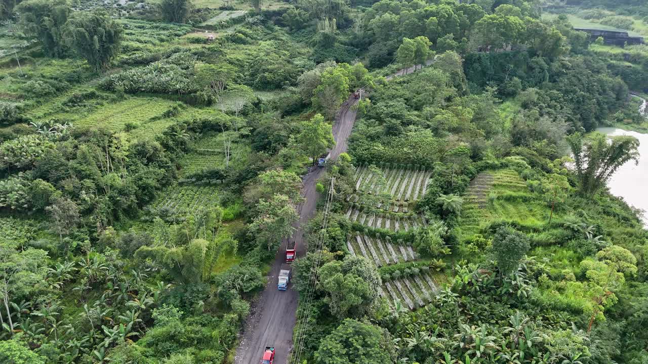 Aerial view of mining trucks driving through the path in the middle of green vegetation of forests and grasslands. Slope of Merapi Volcano, Indonesia.