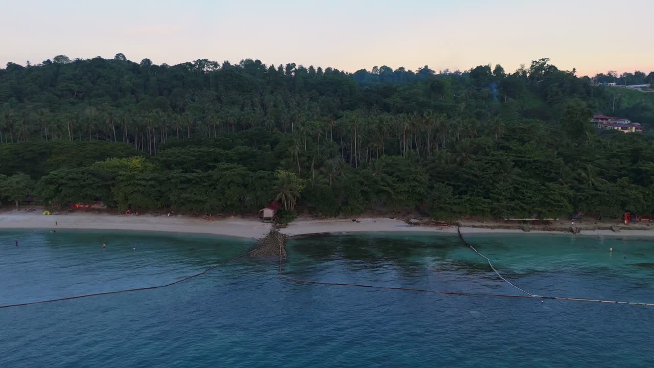 Aerial sunset view of a tropical beach with clear blue water, white sand and dense palm forest. Peaceful coastal atmosphere with evening light highlighting the shoreline and natural island scenery