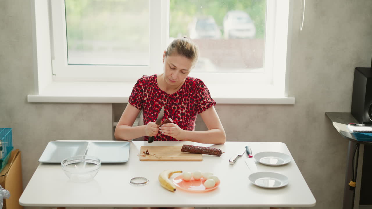 Wide shot of female griller seated by bright window, carefully peeling tough casing from smoked sausage on wooden board, focused expression highlighting precise preparation