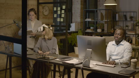 woman emplooyee at desk in the office wearing virtual reality glasses while her coworker is controlling her with tablet