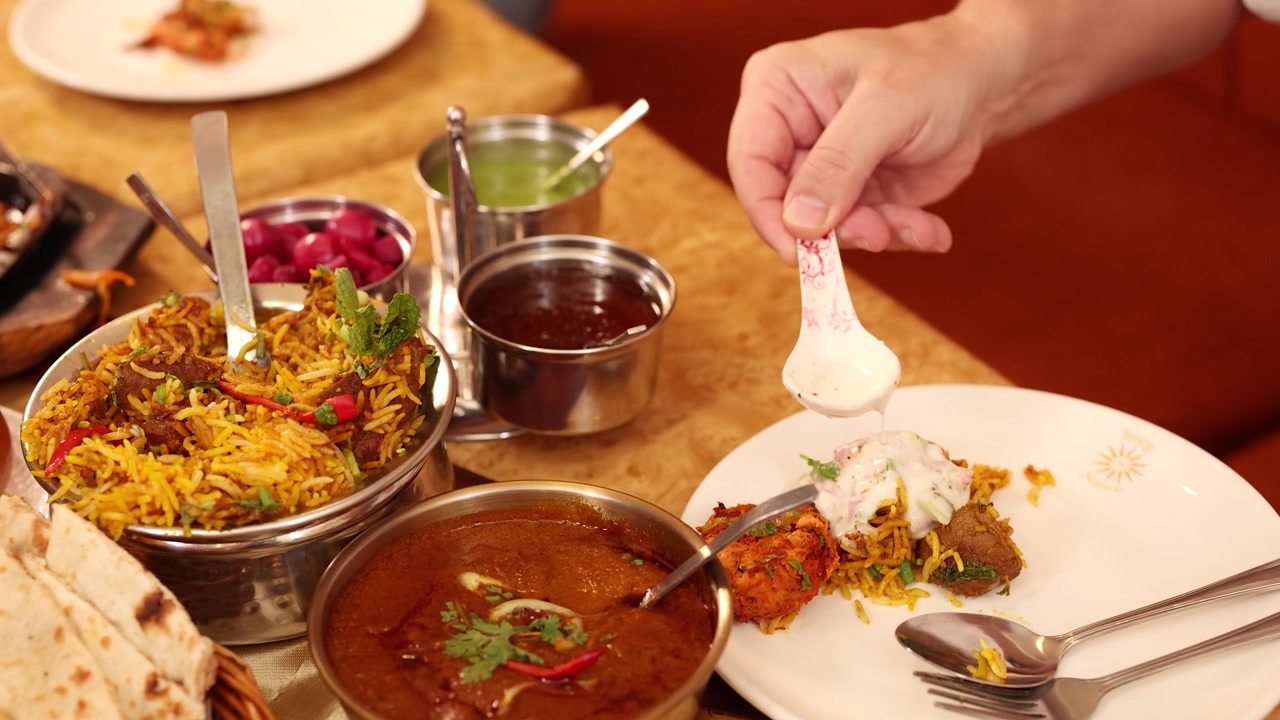 Nærbillede af en hånd, der hælder hvid sauce over biryani-ris blandt forskellige indiske retter og karryretter.