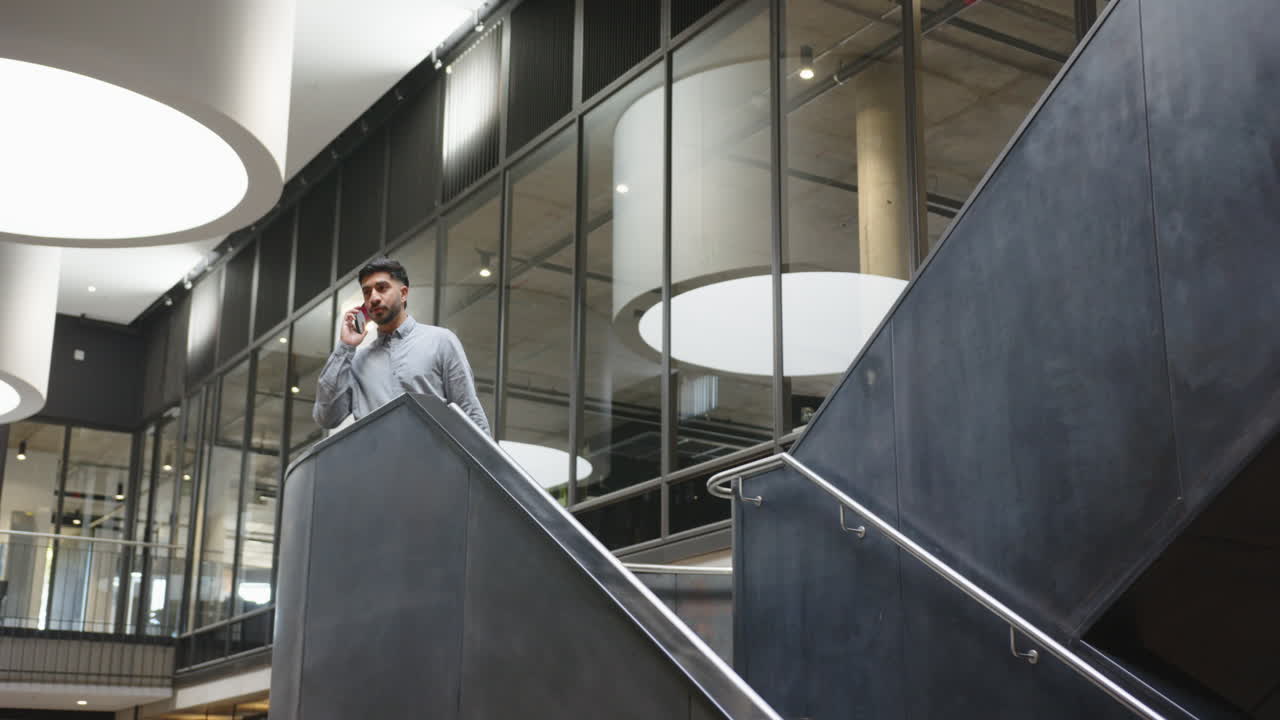 Talking on smartphone, businessman standing on modern office staircase looking serious
