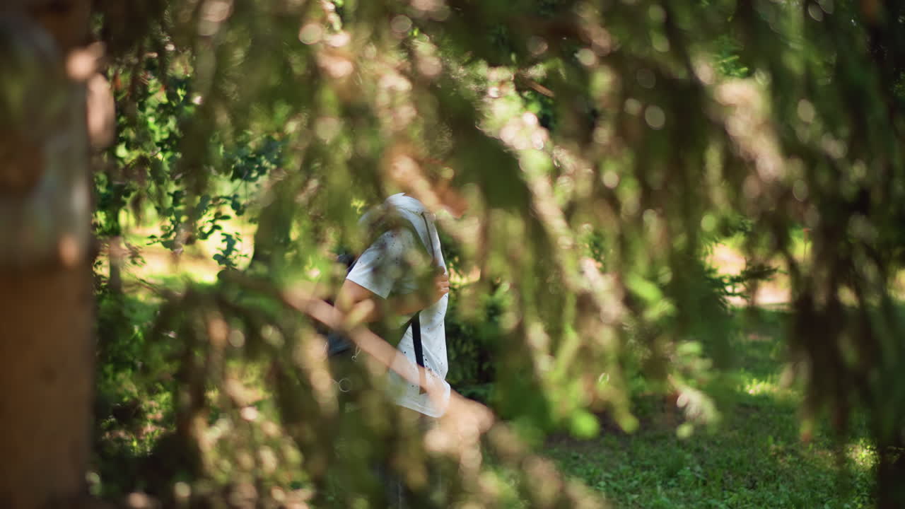 배낭을 메고 전나무 그늘을 걷는 젊은 사람, 옆모습으로 걷고 있으며, 나뭇가지 사이로 카메라가 살짝 보이고, 땅에는 얼룩덜룩한 햇빛이 비치며, 조용한 숲 속에서 사색에 잠긴 야외 풍경