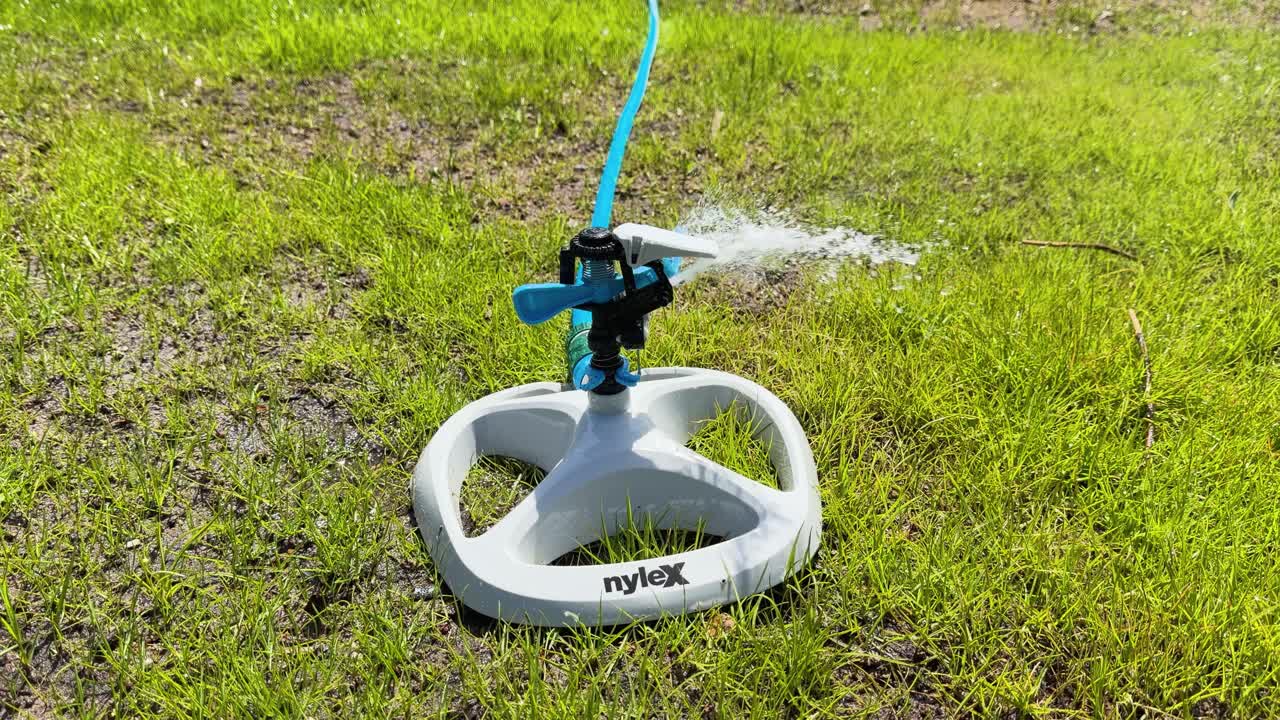 Slow motion rotating garden sprinkler sprays water across bright green lawn under sunlight