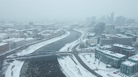 drone shot of river neris winds gracefully through vilnius, its icy banks blanketed in snow as a gentle blizzard falls from the grey sky