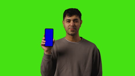 portrait of young man holding blue screen mobile phone to camera standing against green screen 1