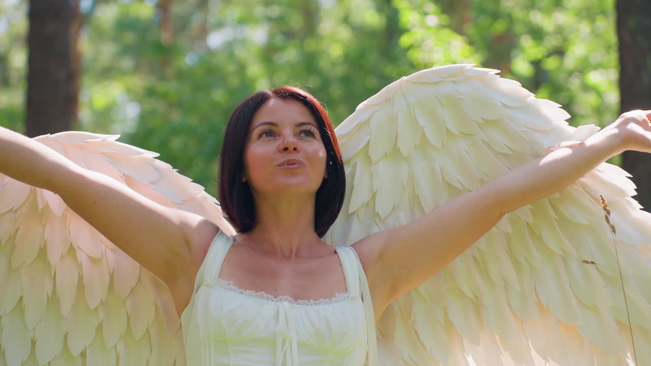 Goddess of love with radiant angel wings lifts arms gracefully toward glowing sunlight in green forest, surrounded by warm light and soft leaves creating peaceful ethereal divine magical atmosphere