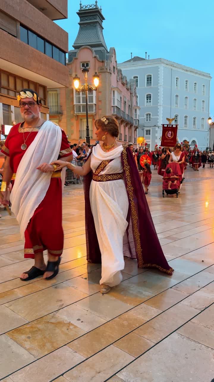 Madrid, Spagna, 8 agosto 2025: Coppia vestita da nobili romani durante una sfilata storica serale. Uomo e donna con tuniche e toghe romane tradizionali che camminano su una piazza pavimentata al tramonto