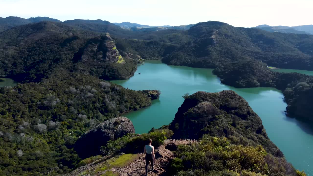 Drone view of young caucasian woman walking on top of a rock with a view of a bay and islands with mountains, greenery and turquoise water on a sunny day in Duke's Nose, Northland, New Zealand.