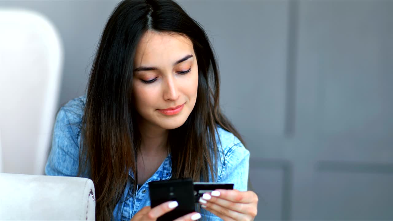 joven bonita comprando en línea con tarjeta de crédito y teléfono inteligente acostado en un sofá en la sala de estar en casa