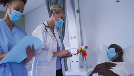 Diverse-female-doctors-talking-to-male-patient-in-hospital-bed-all-wearing-face-masks