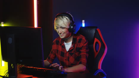 young woman with short blond hair in big headphones sitting in the gamer chair in front of the computer screen and celebrating that she has won the game