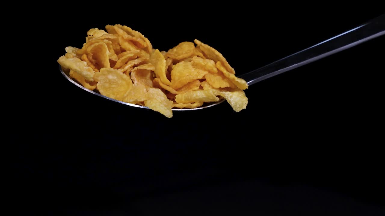A spoon releases corn cereal flakes, captured mid-air against a dark backdrop, highlighting motion and texture