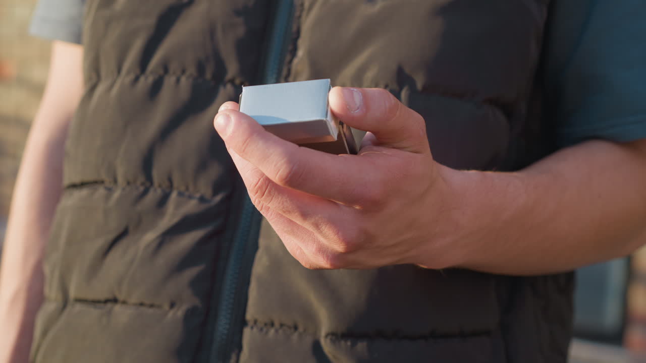 partial view of person wearing padded vest holding cigarette pack in one hand, slowly closing lid while standing outdoors near brick building during daylight