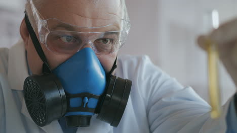 a scientist at a pharmaceutical company developing drugs wearing a blue respirator and safety glasses holds a new drug a glass tube with a yellow liquid in his hands and looks at it. high quality 4k footage