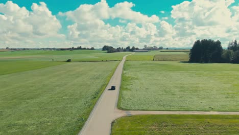 a single car driving on a long, winding road through vibrant green fields under a partly cloudy sky, daytime, aerial view