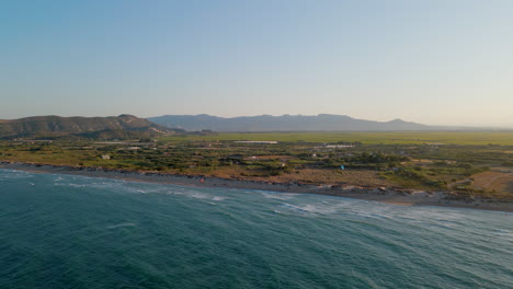 Vista-Aérea-De-La-Costa-En-La-Playa-De-Mareny-Con-Los-Campos-De-Arroz-Y-Las-Montañas-Al-Fondo,-Valencia,-España