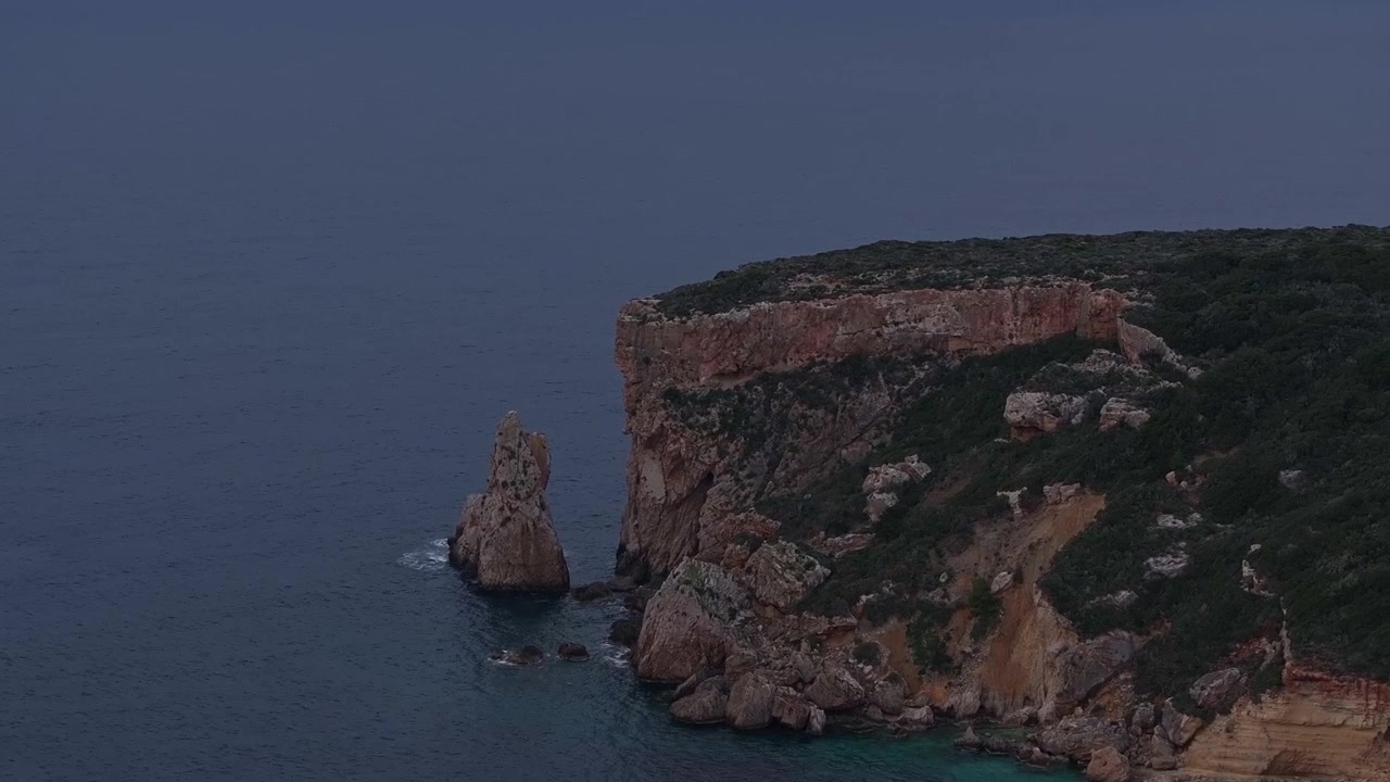 Telefoto luftbillede af dramatiske stejle kystklipper og en isoleret havstabel stående i turkist vand nær Pylos, Grækenland, under en mørk, stemningsfuld aftenshimmel i skumringen.