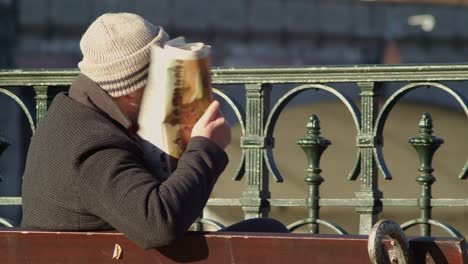 a man on a bench folds a newspaper and takes off his glasses