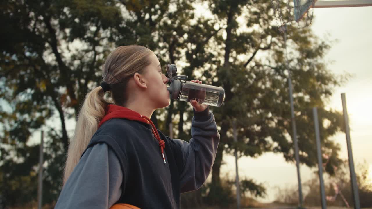fotografía en primer plano de una vista trasera de una chica rubia con un peinado de cola de caballo bebiendo agua de una botella deportiva durante su práctica de baloncesto matutino y mirando el amanecer en verano