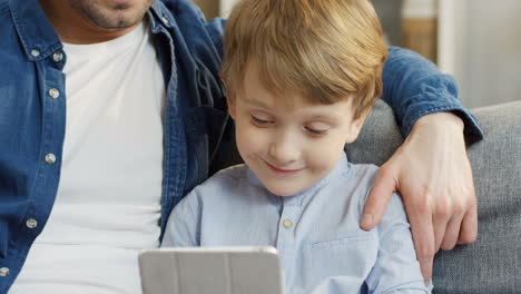 Close-Up-Of-The-Attractive-Father-Sitting-On-The-Couch-And-Hugging-His-Small-Son-While-They-Watching-Something-On-The-Tablet-Device