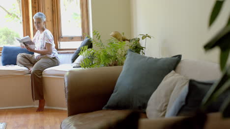 Happy-senior-biracial-woman-on-couch-reading-book-at-window-at-home,-slow-motion