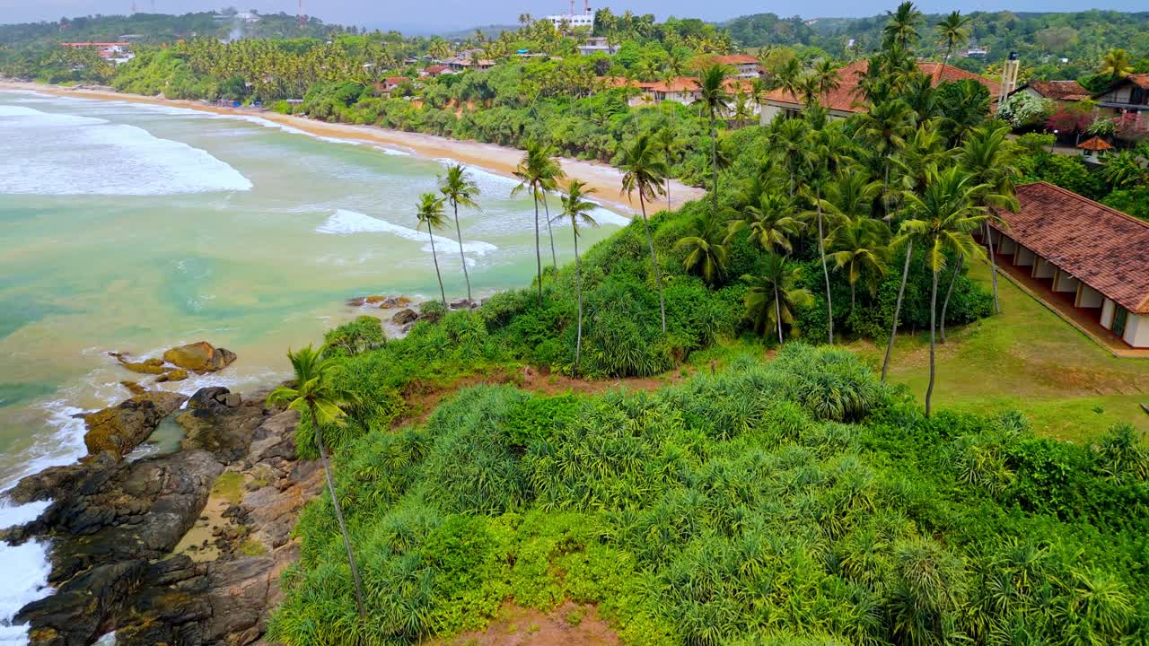 An aerial drone view of SK Town beach in Weligama, Sri Lanka, showcasing golden sand, gentle waves, and a lush line of palm trees along the shore.