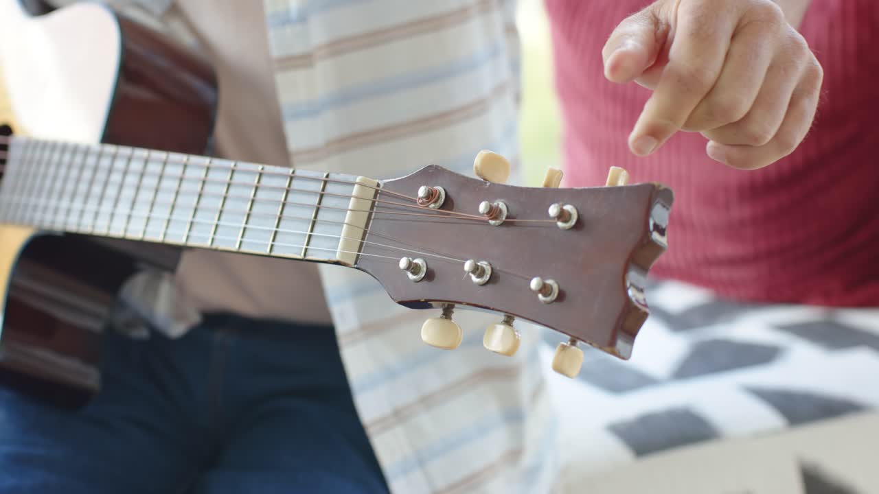 Right hand is reaching, turning guitar tuners on couch while partner holding guitar, checking pitch
