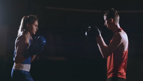 Schöne-Brünette-Boxerin-Trainiert-Mit-Einem-Trainer-In-Einem-Dunklen-Raum-Schläge-Auf-Die-Pfoten