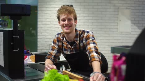 Portrait-of-handsome-young-man-working-at-cash-register-in-supermarket,-posing-looking-at-camera-and-smiling-happily.-Sitting-at-the-cash-register