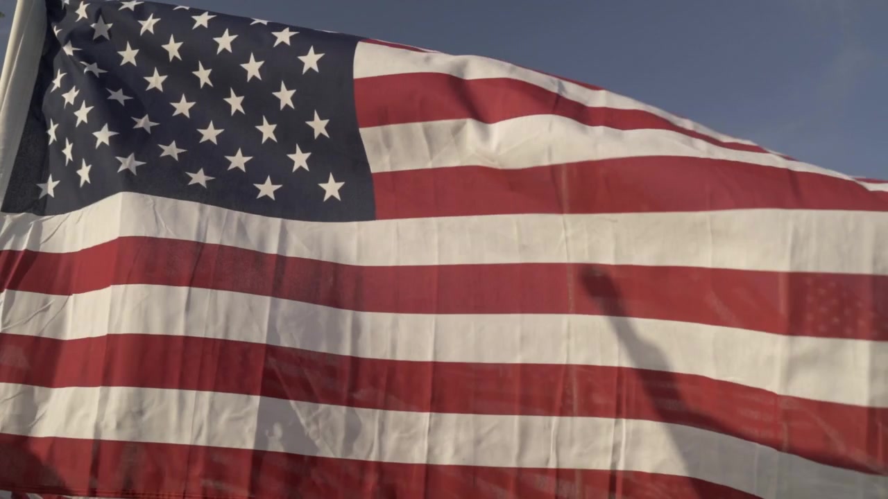 Flere amerikanske flagg vaier i vinden i en utendørs minnepark, noe som skaper en mektig og patriotisk scene. Den lyse himmelen fremhever flaggenes røde, hvite og blå farger.