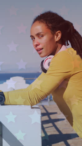 múltiples iconos de estrellas contra una mujer afroamericana que usa un teléfono inteligente en el paseo marítimo