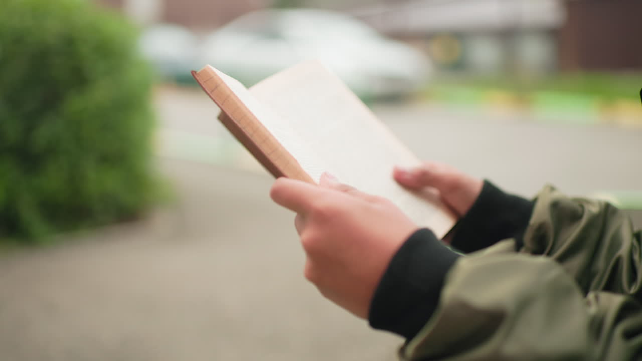 Close up of hands holding and flipping through pages of book outdoors with blurred background showing concentration, study habit, and pursuit of knowledge