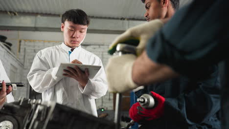 mechanic demonstrates tool usage on engine while students take notes on tablets, second mechanic observes student's input, industrial automotive workshop filled with tools, machinery in background